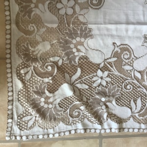 May include: A white tablecloth with a beige lace border featuring a floral pattern. The lace design includes intricate swirls and leaves.