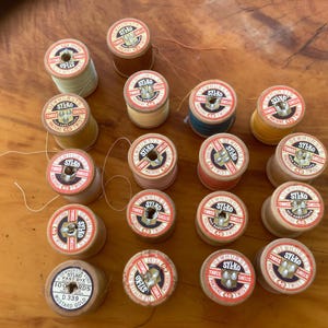 Può includere: Una collezione di rocchetti di filo Sylko vintage in vari colori, tra cui giallo, verde e rosso. Ogni rocchetto ha un'etichetta circolare con il logo e il testo Sylko. I rocchetti sono disposti su una superficie di legno.