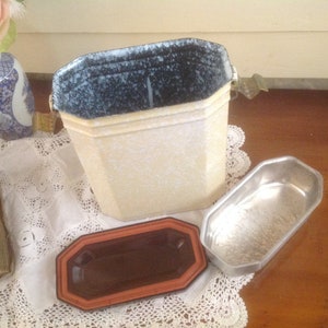 May include: A vintage white enamelware container with a speckled blue interior and a metal lid. The container has a rectangular shape with rounded corners and a small metal handle on the side. The container is sitting on a white lace tablecloth.