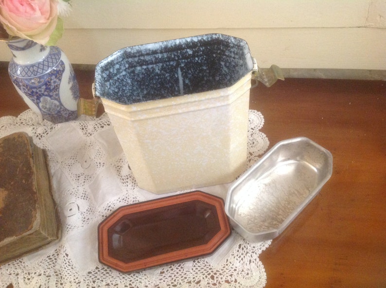 May include: A vintage white enamelware container with a speckled blue interior and a metal lid. The container has a rectangular shape with rounded corners and a small metal handle on the side. The container is sitting on a white lace tablecloth.