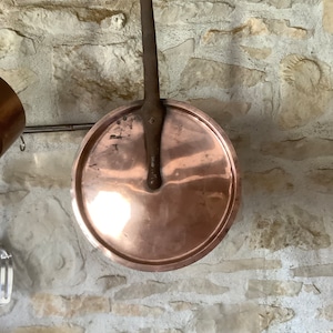 Op de afbeelding: Een koperen pandeksel met een lange, donkerbruine handgreep die aan een haak hangt. Het deksel is rond en heeft een glanzend, reflecterend oppervlak. De achtergrond is een lichtgrijze stenen muur.