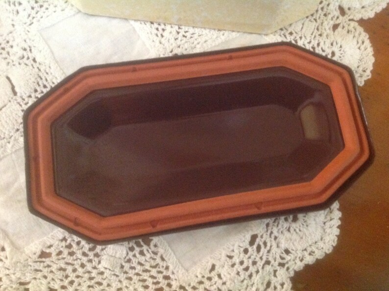 May include: A terracotta-colored rectangular dish with a slightly rounded edge. The dish has a dark brown glaze inside and a smooth, glossy finish.