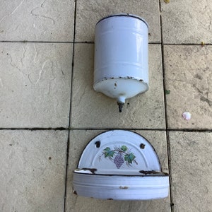 Puede incluir: Dos lavabos de pared vintage de esmalte blanco. El lavabo superior es cilíndrico y el lavabo inferior es un semicírculo con un diseño de vid.