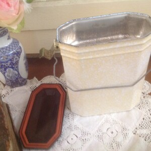May include: A white metal container with a silver interior and a metal handle. The container has a speckled white finish and is sitting on a white lace tablecloth.