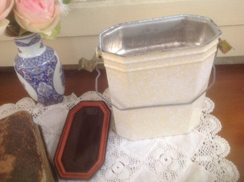 May include: A white metal container with a silver interior and a metal handle. The container has a speckled white finish and is sitting on a white lace tablecloth.