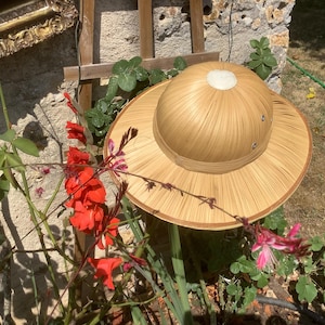Può includere: Un cappello safari marrone chiaro con tesa larga e corona arrotondata. Il cappello è realizzato in materiale intrecciato e ha un bordo marrone scuro. Il cappello è davanti a un muro di pietra con fiori rossi.