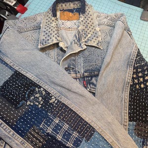May include: A light-wash denim jacket featuring intricate sashiko-style white stitching patterns on the collar and shoulders. The sleeves and lower sections are adorned with various dark blue and indigo fabric patches, showcasing diverse geometric and floral patterns.