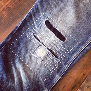 May include: Close-up of a pair of blue denim jeans with a large, rectangular patch sewn over a hole in the knee. The patch is stitched with white thread in a decorative pattern.