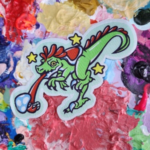 May include: Green dinosaur with red and white accents, holding a blue and white egg in its mouth. The dinosaur is surrounded by yellow stars on a light blue background.