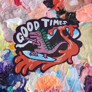 May include: A pink and green dinosaur surfing on a wave with the text "GOOD TIMES" in white letters on a black background.
