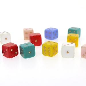 Puede incluir: Un conjunto de dados coloridos en varios tonos, incluyendo rojo, azul, verde y amarillo. Cada dado tiene puntos dorados que indican los números. Los dados están dispuestos sobre una superficie blanca.