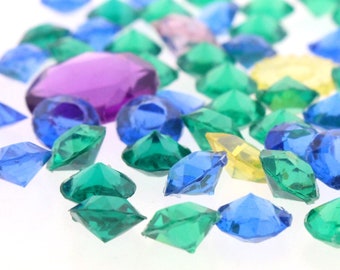 Puede incluir: Surtido de gemas de plástico de colores en varios tonos de azul, verde y morado. Las gemas están esparcidas sobre una superficie blanca.