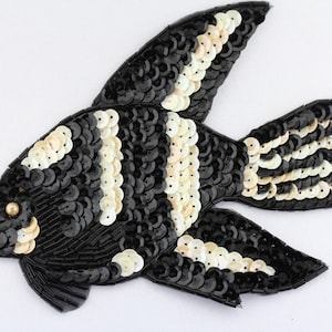 May include: A decorative fish applique, featuring a black and white sequin design. The fish has a detailed pattern with alternating stripes of black and white sequins, and a small gold bead for the eye. Ideal for embellishing clothing or accessories.