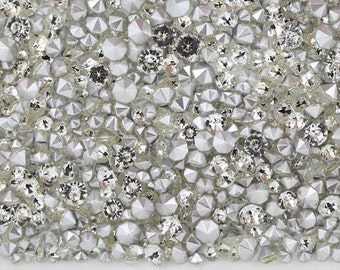 May include: Close-up of a collection of sparkling, silver-colored rhinestones. The rhinestones vary in size and shape, with faceted and rounded designs. The arrangement creates a shimmering, textured surface, ideal for embellishments and crafts.