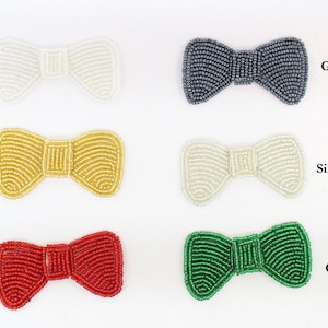 Puede incluir: Seis apliques de lazo de cuentas en varios colores: blanco (Iris), plomo, dorado, plateado, rojo y verde. Cada lazo está hecho de pequeñas cuentas y tiene una forma clásica. Los nombres de los colores están escritos junto a cada lazo.