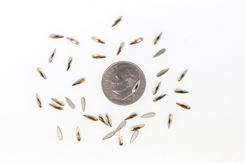 May include: A scattering of silver teardrop-shaped rhinestones, some with a gold hue, are arranged around a US dime coin. The rhinestones are approximately the same size as the coin.