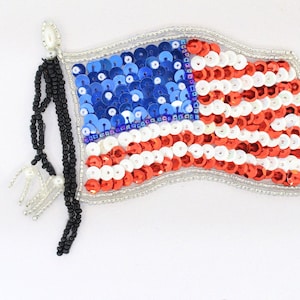 May include: A red, white, and blue American flag patch made of sequins. The flag is sewn onto a white fabric background. The flag has a black beaded tassel attached to the top.