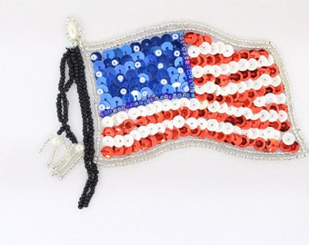 May include: A red, white and blue American flag patch made of sequins. The flag is sewn onto a white fabric background. The flag has a black beaded tassel attached to the top.