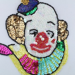 Könnte beinhalten: Eine bunte Pailletten-Clown-Gesichtsapplikation mit weißem Gesicht, schwarzen Augen, roten Lippen und einer roten Nase. Der Clown trägt einen grün-blauen Hut und einen gelb-rosa-blauen Kragen.