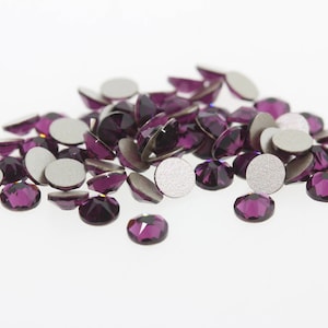 May include: A pile of purple rhinestones with silver backing. The rhinestones are scattered on a white surface.