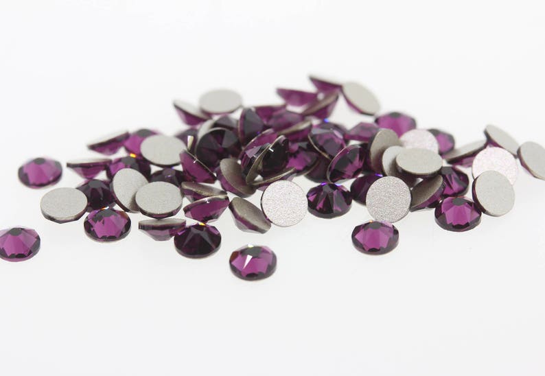 May include: A pile of purple rhinestones with silver backing. The rhinestones are scattered on a white surface.