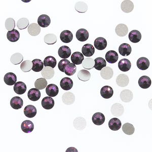 May include: A scattering of small, round, purple rhinestones on a white background. The rhinestones are scattered randomly and some are overlapping.