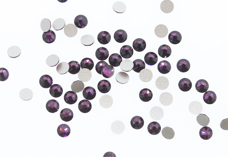 May include: A scattering of small, round, purple rhinestones on a white background. The rhinestones are scattered randomly and some are overlapping.