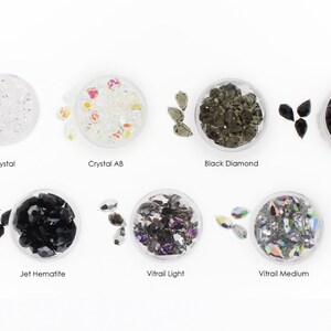 May include: Assortment of clear, black, and iridescent glass rhinestones in various shapes and sizes. The rhinestones are displayed in small clear containers with labels indicating their color and name: Crystal, Crystal AB, Black Diamond, Jet, Jet Hematite, Vitrail Light, and Vitrail Medium.