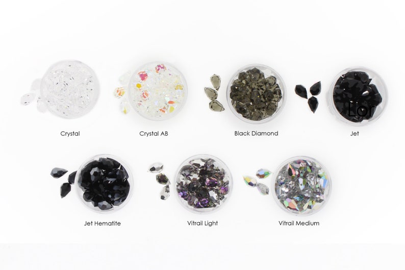May include: Assortment of clear, black, and iridescent glass rhinestones in various shapes and sizes. The rhinestones are displayed in small clear containers with labels indicating their color and name: Crystal, Crystal AB, Black Diamond, Jet, Jet Hematite, Vitrail Light, and Vitrail Medium.