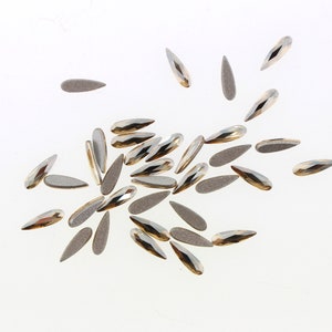 May include: A scattering of silver and gold teardrop-shaped nail rhinestones on a white background.
