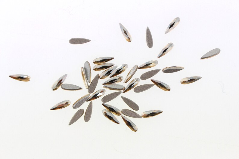 May include: A scattering of silver and gold teardrop-shaped nail rhinestones on a white background.