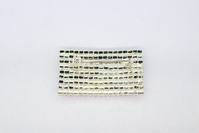 Puede incluir: Un broche plateado de forma rectangular con un pasador en la parte posterior. El broche est&aacute; hecho de peque&ntilde;os diamantes de imitaci&oacute;n cuadrados.