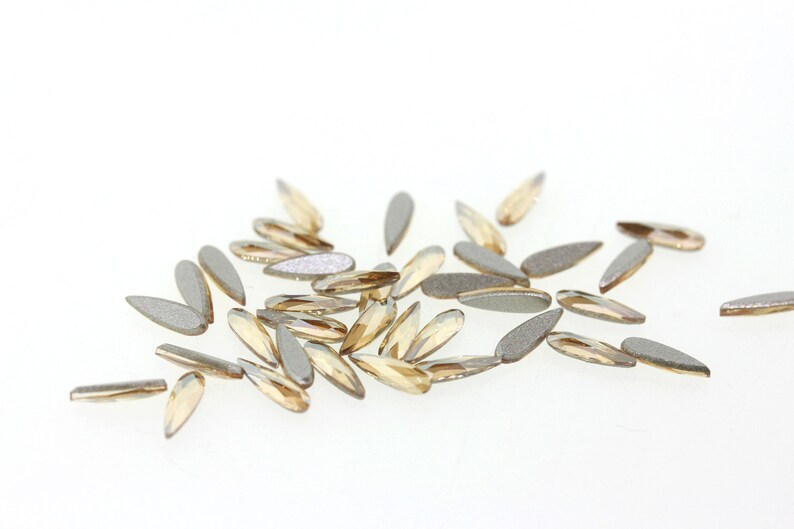 May include: A scattering of small, teardrop-shaped rhinestones in a light gold color. The rhinestones have a slightly iridescent finish.