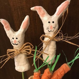 Può includere: Due figurine di coniglio in ceramica bianca con orecchie rosa e fiocchi di paglia marroni, in piedi in un nido di ceramica con tre carote arancioni.