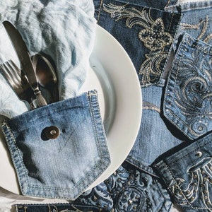 Puede incluir: Un plato blanco con una servilleta de mezclilla azul y cubiertos metidos en un bolsillo de mezclilla. La servilleta est&aacute; doblada y los cubiertos est&aacute;n colocados dentro del bolsillo. El plato est&aacute; rodeado de tela de mezclilla con dise&ntilde;os bordados.