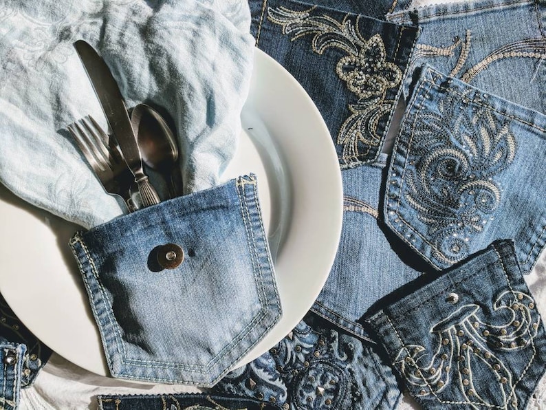 Puede incluir: Un plato blanco con una servilleta de mezclilla azul y cubiertos metidos en un bolsillo de mezclilla. La servilleta est&aacute; doblada y los cubiertos est&aacute;n colocados dentro del bolsillo. El plato est&aacute; rodeado de tela de mezclilla con dise&ntilde;os bordados.
