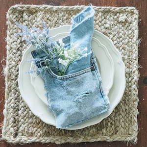 May include: A white plate with a blue denim napkin folded inside. The napkin has a pocket with a sprig of white and purple flowers inside. The plate is on a woven placemat.