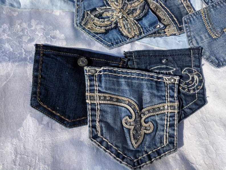Puede incluir: Tres bolsillos de mezclilla con costuras y adornos decorativos. Los bolsillos son azules y tienen una variedad de dise&ntilde;os, que incluyen una flor de lis, una flor y una luna creciente.