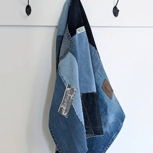 May include: A patchwork denim towel hanging on a white wall. The towel is made of various shades of blue denim, with visible seams and patches. A brown leather patch is sewn on the towel. The towel is hanging from a black hook.