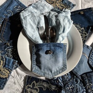 Puede incluir: Un plato blanco con una servilleta de lino azul y cubiertos dentro de un bolsillo de mezclilla. El plato est&aacute; rodeado de tela vaquera con dise&ntilde;os bordados.