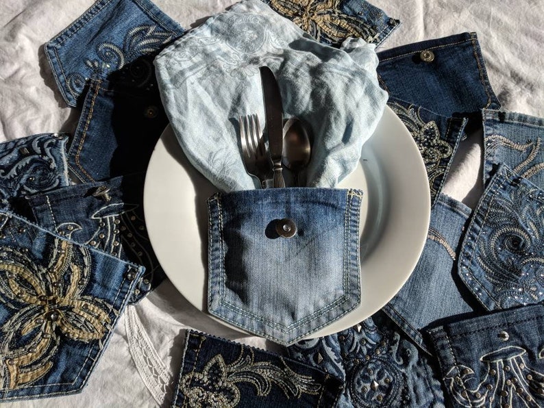 Puede incluir: Un plato blanco con una servilleta de lino azul y cubiertos dentro de un bolsillo de mezclilla. El plato est&aacute; rodeado de tela vaquera con dise&ntilde;os bordados.