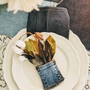 May include: A white plate with a smaller white plate on top. A denim pocket holds a bouquet of brown feathers and green leaves with white cotton balls.