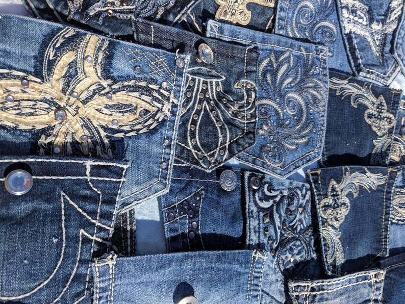 Puede incluir: Un primer plano de una tela de mezclilla de retazos con varios dise&ntilde;os bordados. La tela presenta intrincados patrones en hilo dorado y negro, con algunos dise&ntilde;os adornados con pedrer&iacute;a. La mezclilla es de color azul oscuro y tiene un aspecto desgastado.