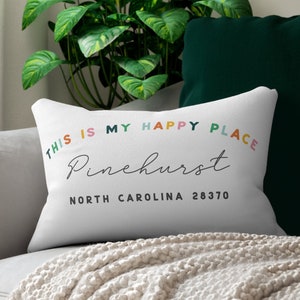 May include: White rectangular cushion with the text "THIS IS MY HAPPY PLACE" in colourful letters. Below, the word "Pinehurst" is written in cursive, followed by "NORTH CAROLINA 28370". The cushion is on a grey sofa.