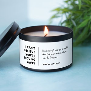 May include: Black candle with a white label that reads "I can't believe you're moving away". The label also includes a message that says "We are going to miss you so much! Good luck on the next adventure. Love, The Thompsons". The candle is on a light blue surface.