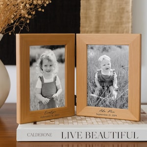 May include: A hinged double photo frame made of light-toned wood. Each frame holds a black and white photograph of a child. The left frame is engraved with the name Evelyn and February 16, 2022. The right frame features Ada Mae and September 19, 2024.