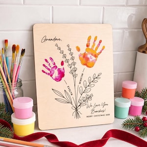 May include: A rectangular wooden plaque featuring a black line art illustration of flower stems. Two colorful handprints in pink and orange serve as flower blooms. The text reads Grandma, Evelyn, Winston, We Love You Bunches! MERRY CHRISTMAS 2026.