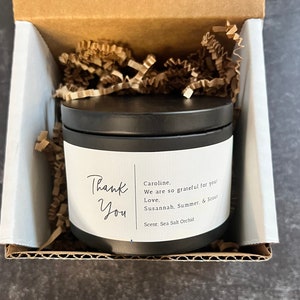 May include: A black candle jar with a white label that says "Thank You" and includes a personalized message. The candle is in a cardboard box with brown packing paper.