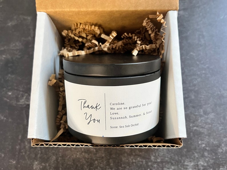 May include: A black candle jar with a white label that says "Thank You" and includes a personalized message. The candle is in a cardboard box with brown packing paper.