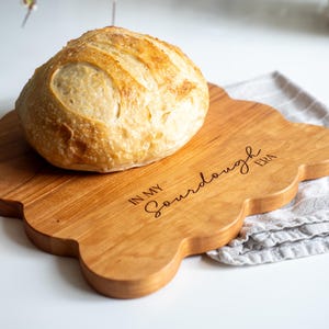May include: A wooden cutting board with a scalloped edge, engraved with the text "In My Sourdough Era". A loaf of sourdough bread sits on the board.